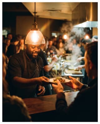 A chef handing a small tasting plate across a dark counter.